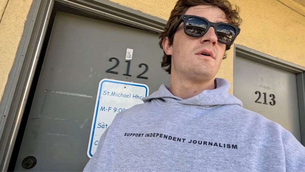 Shirley wearing sunglasses and a gray hoodie reading "SUPPORT INDEPENDENT JOURNALISM" standing in front of doors numbered 212 and 213.