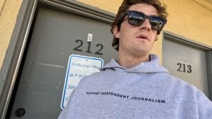 Shirley wearing sunglasses and a gray hoodie reading "SUPPORT INDEPENDENT JOURNALISM" standing in front of doors numbered 212 and 213.