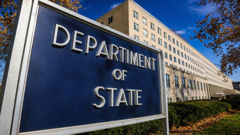 Blue sign reading "DEPARTMENT OF STATE" in front of a large beige government office building on a sunny day