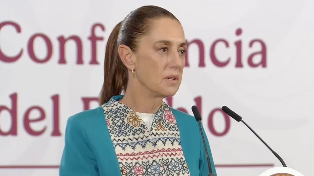 Sheinbaum with pulled-back brown hair and gold hoop earring speaking at a podium, wearing a teal jacket over a patterned blouse, with blurred conference text behind her