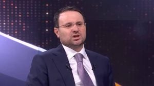 Justice Minister Akın Gürlek in suit and tie speaking on a TV panel, wearing glasses and lapel microphones against a dark dotted studio backdrop.