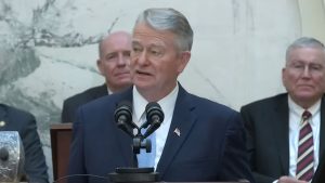 Little in a navy suit and light blue tie speaking at a podium with dual microphones, marble wall and seated men behind him.
