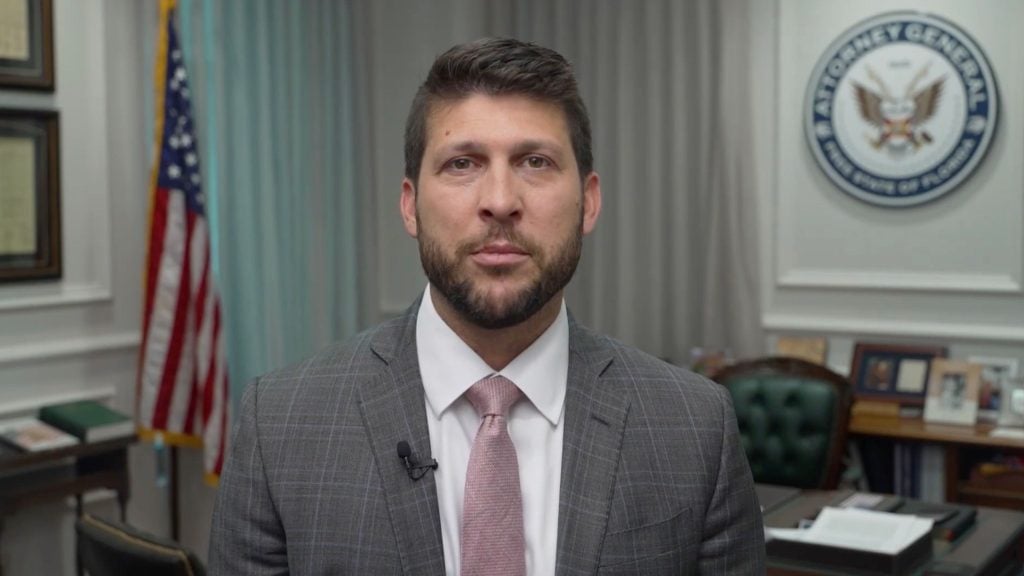 James Uthmeier in a gray plaid suit and pink tie standing in an office with US flag and government seal visible behind him.