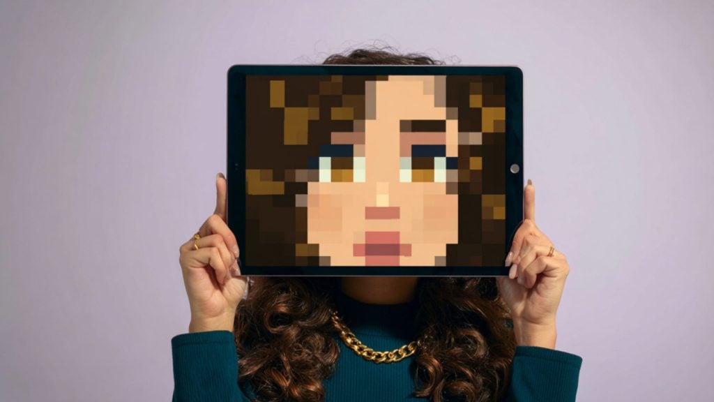 Person holding a tablet in front of their face showing a large pixelated cartoon face, only hair and necklace visible.