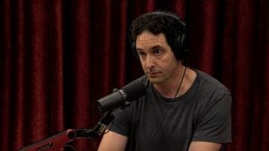 Berenson wearing large black headphones speaking into a studio microphone with red curtains behind him.