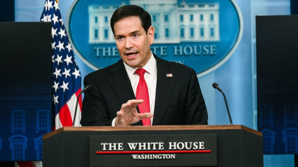 Rubio in dark suit and red tie speaking at a White House podium, gesturing with his hand and an American flag behind him.