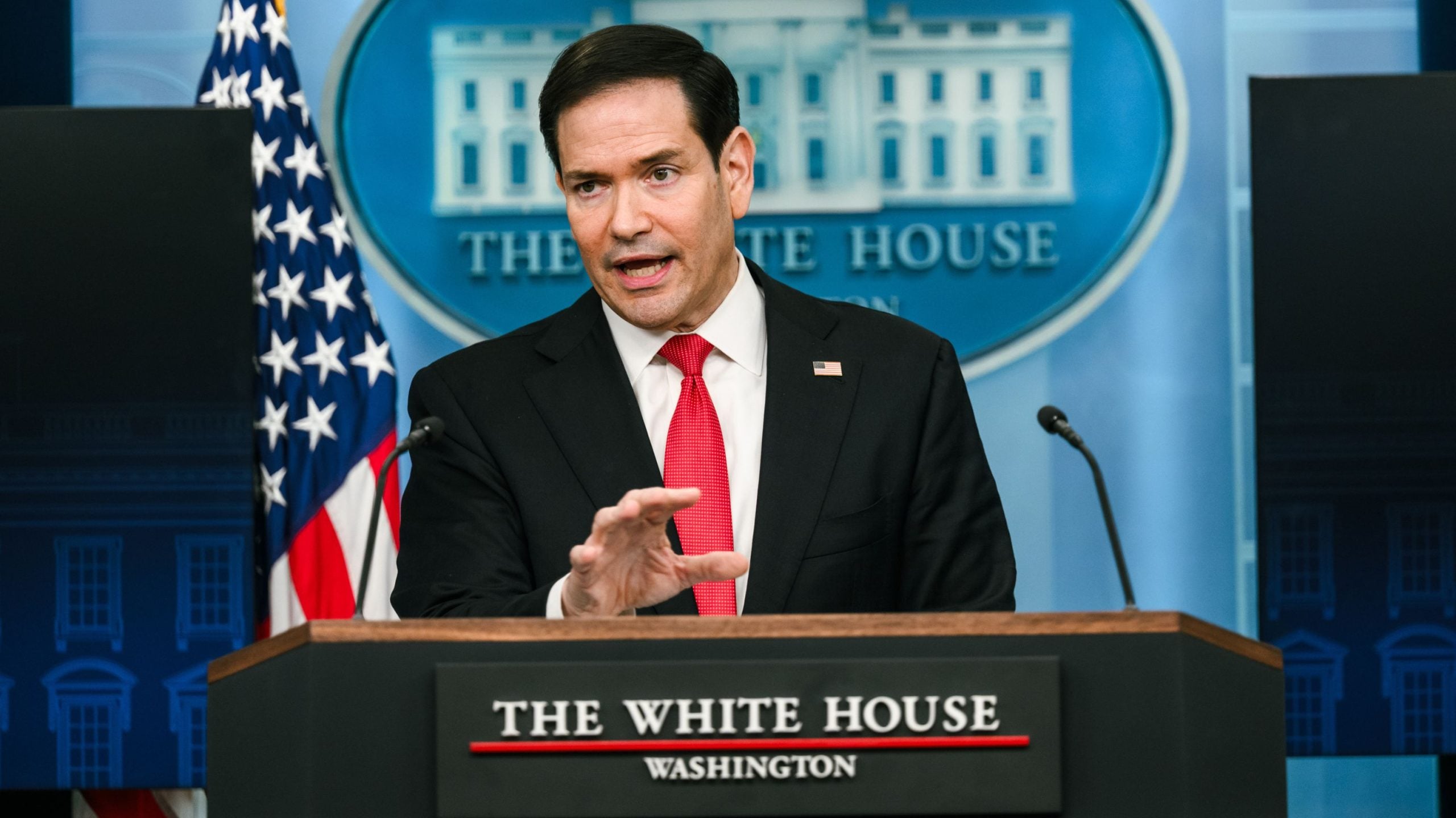 Rubio in dark suit and red tie speaking at a White House podium, gesturing with his hand and an American flag behind him.