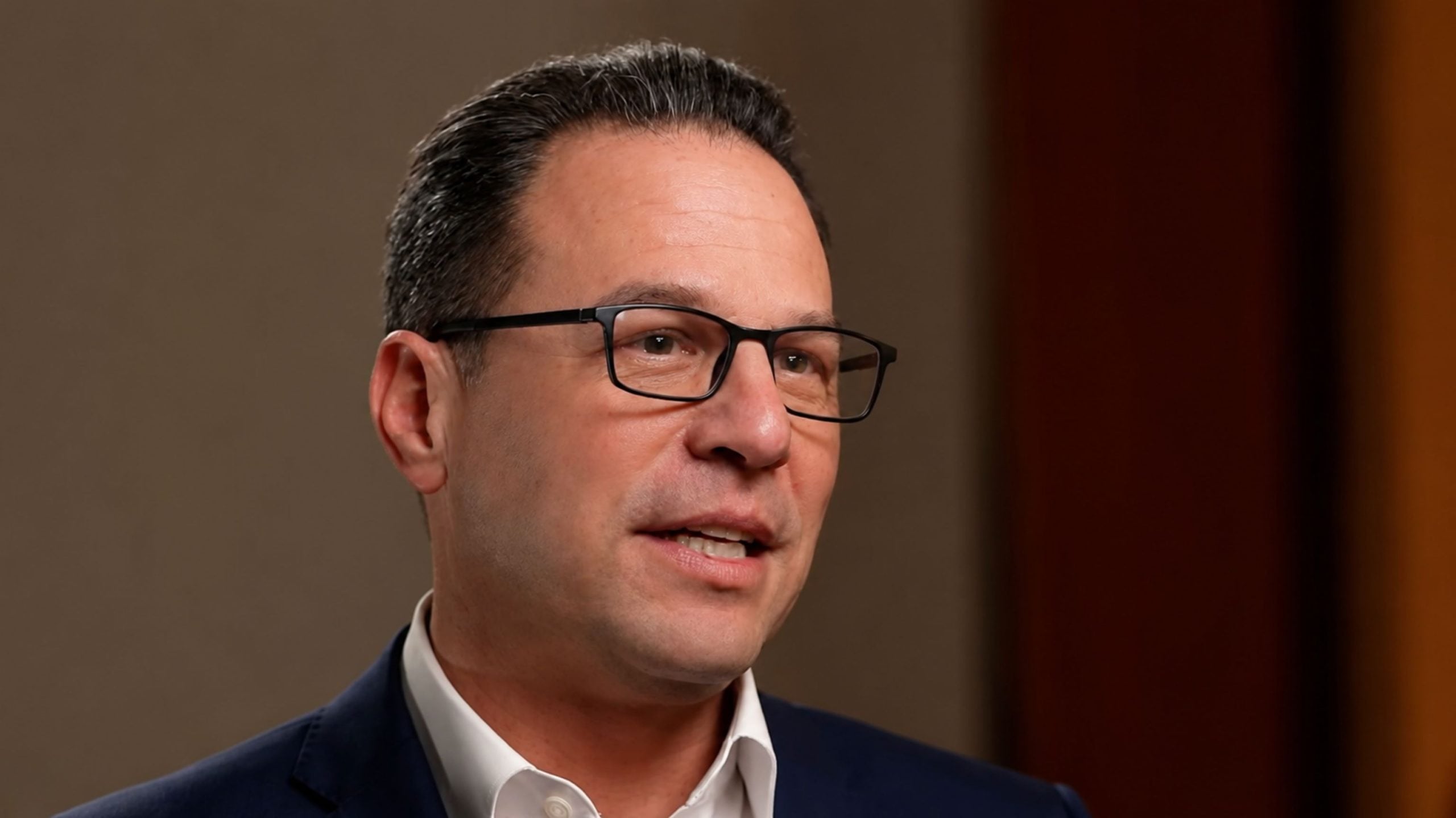 Shapiro wearing black rectangular glasses and a dark suit, speaking in a close-up shot against a blurred brown background.