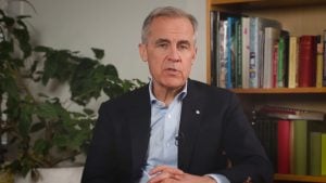 Middle-aged man in a suit jacket and open-collar shirt speaking on camera, seated in front of a bookshelf and a potted plant.